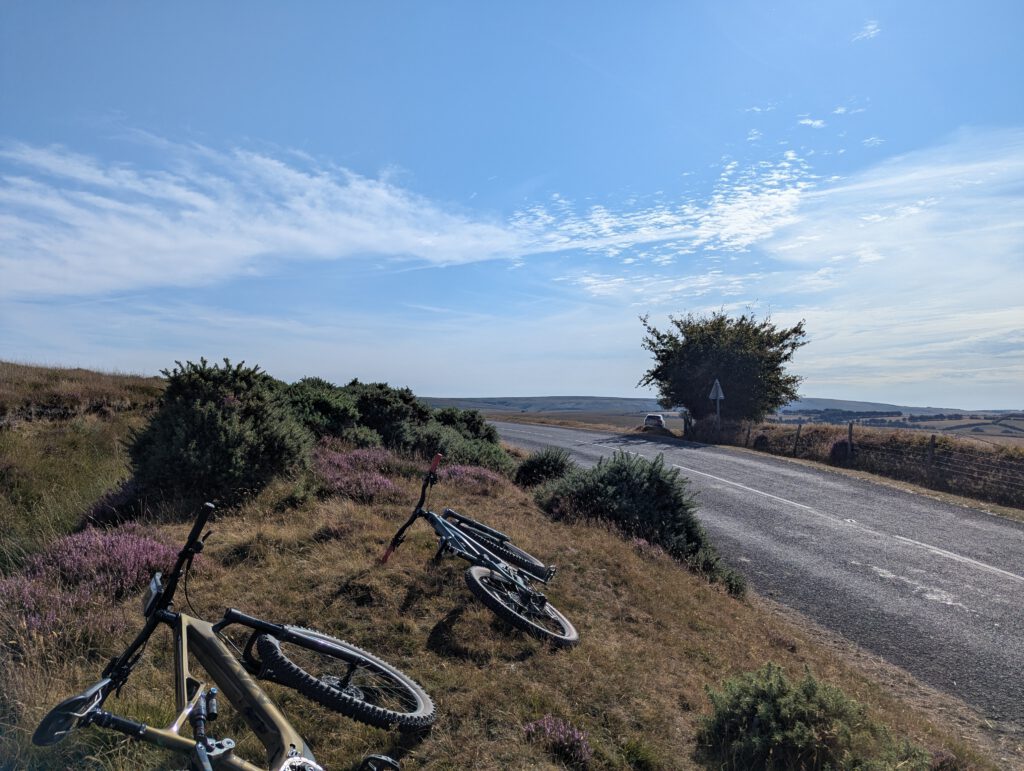 Two mountain bikes laid down in heather beside a quiet moorland road under a wide blue sky