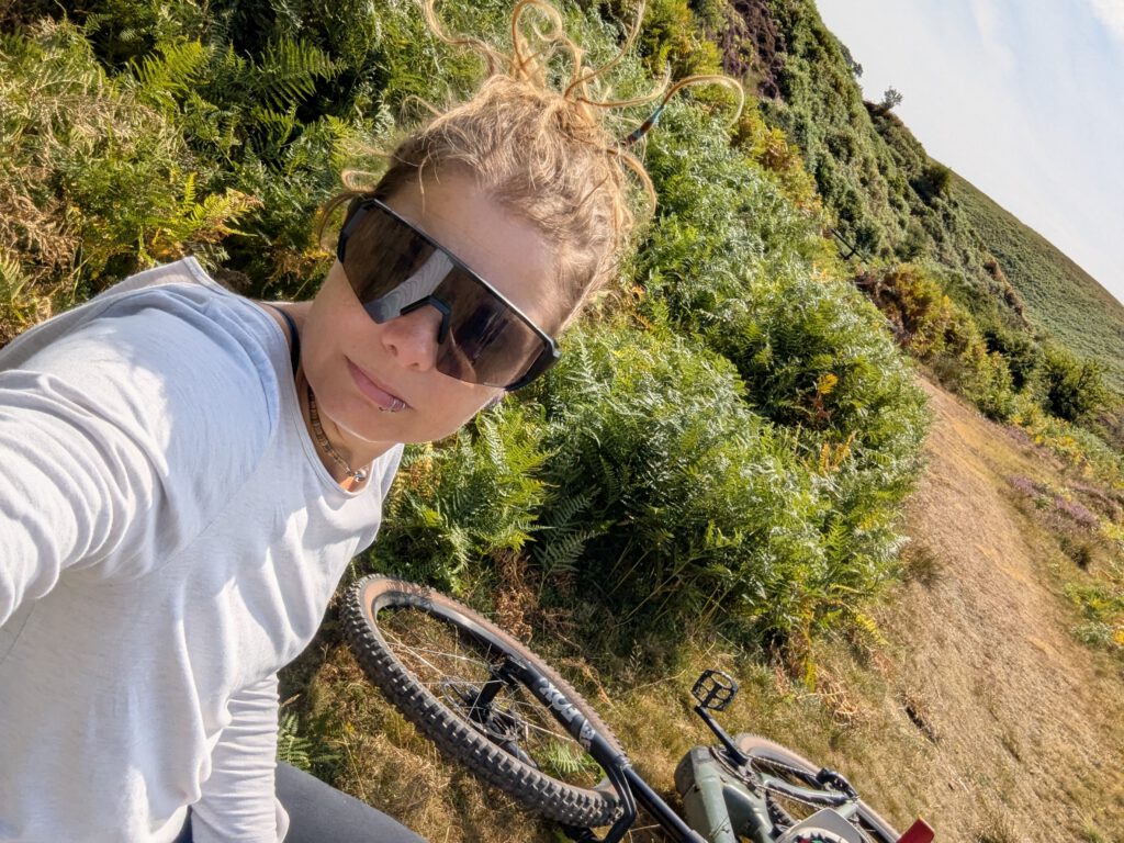 Phe standing on a bracken covered hillside in sunglasses, her mountain bike resting beside her
