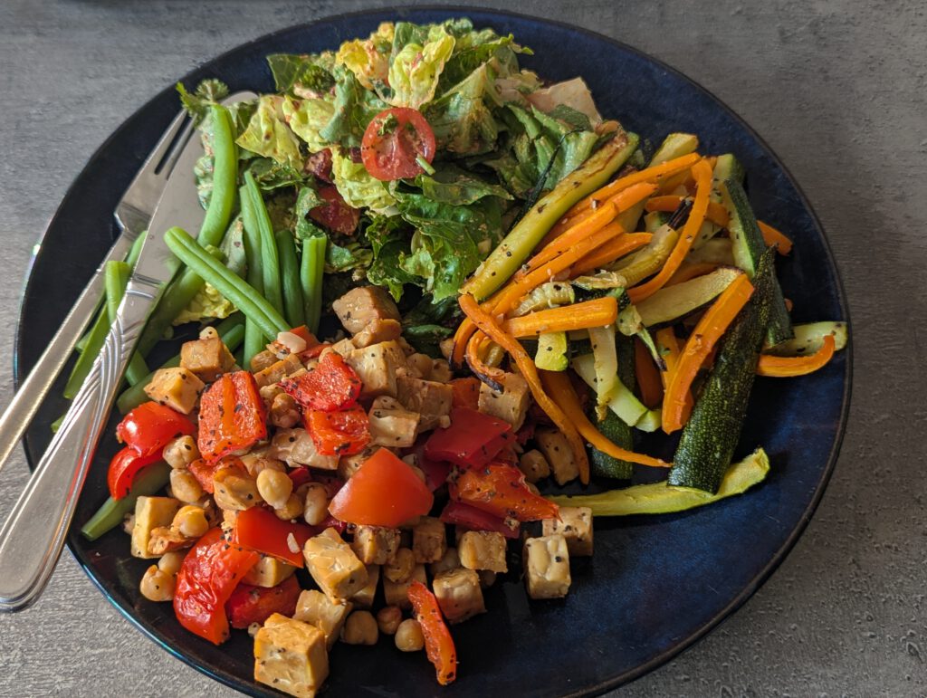 A dark blue plate of tempeh, chickpeas, roasted peppers, courgette, carrots, green beans and salad