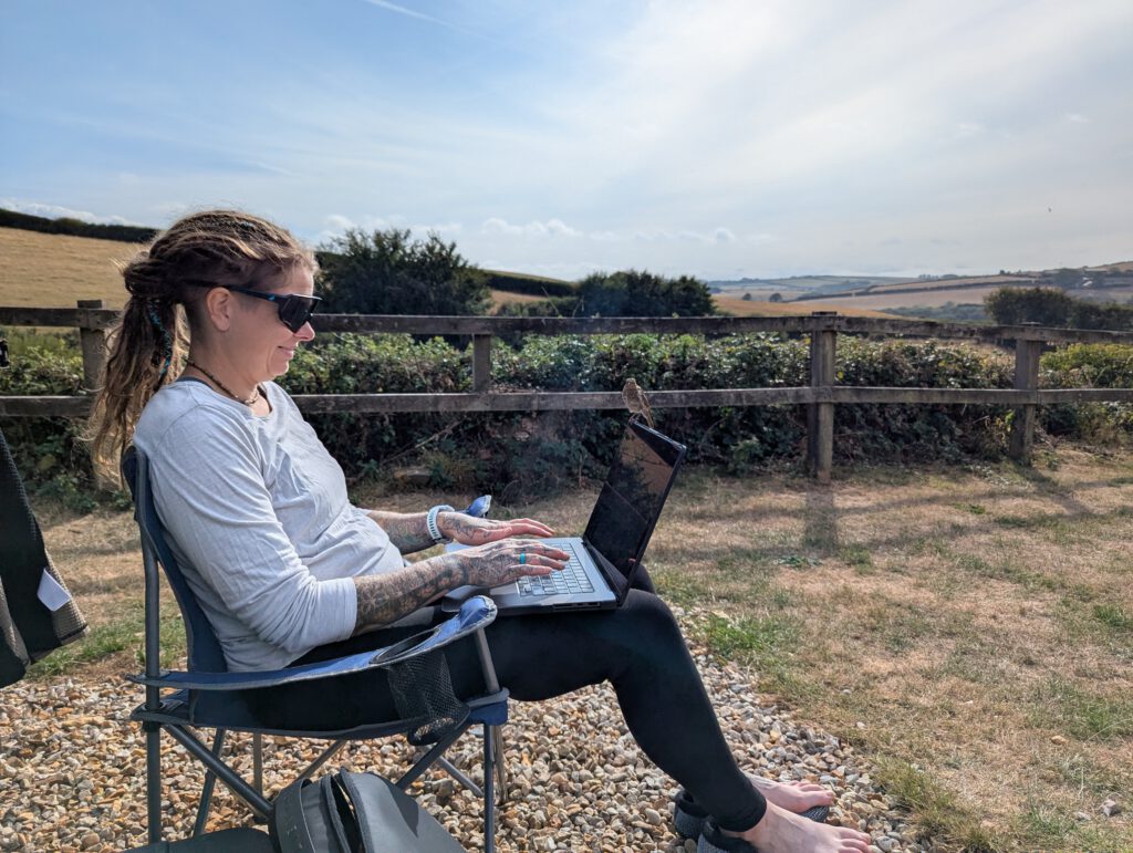 Phe working on a laptop outdoors in a camping chair, with fields and hedgerows behind her